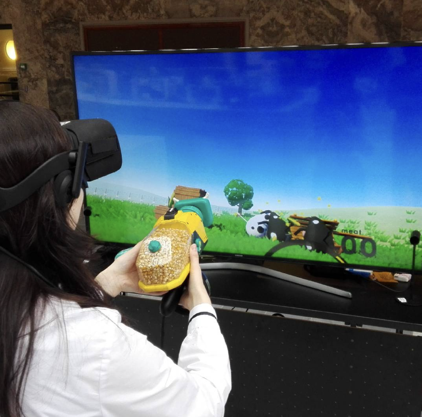 Image of young woman with VR headset holding a yellow and green VR gun that is filled with corn. She is standing for screen with cartoony 3d cows in a field.